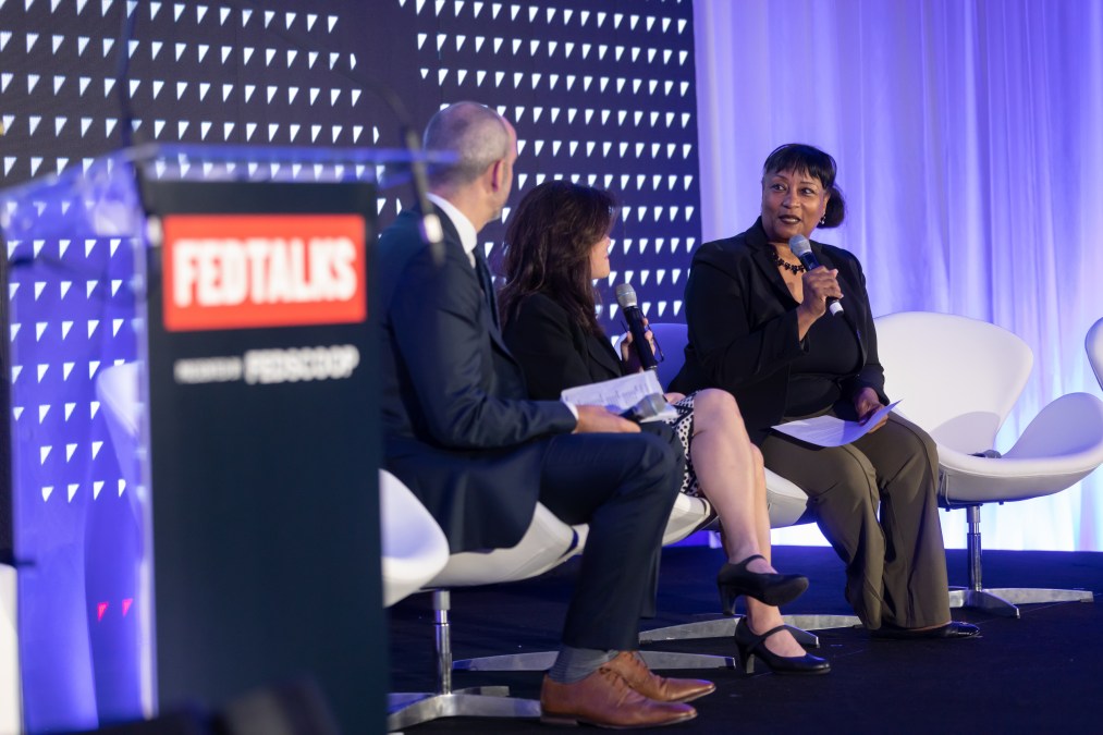 Andrea Brandon sits on a panel with two other people. A lectern with a "FedTalks" sign is in the foreground.