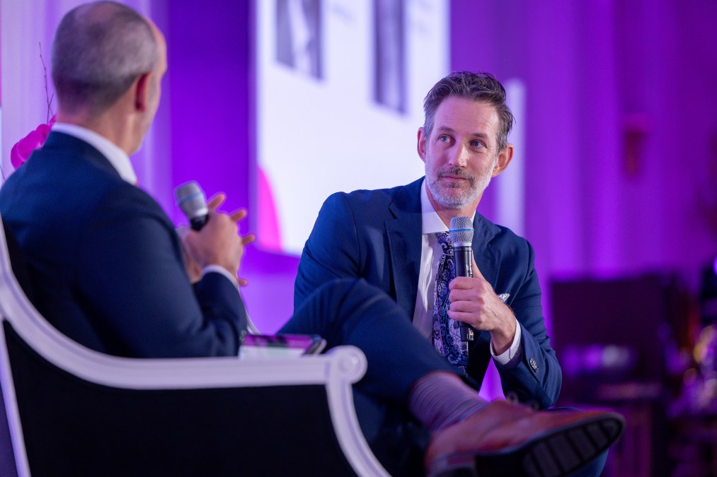 Hoppe is pictured sitting in a chair on stage and holding a microphone. The background is a conference hall lit in pink lights to match the color scheme of the event.