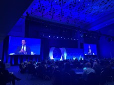 Michael Duffy is pictured speaking onstage in a room at the Washington Convention Center. The stage is illuminated in a deep blue light and he is visible via a livestream on two large screens that flank the stage.