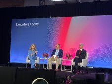 Williams sits in a chair on stage during the panel. The moderator and his fellow panelist sit to the left. They are in front of a multicolor backdrop that says "Executive Forum" at the top left corner.
